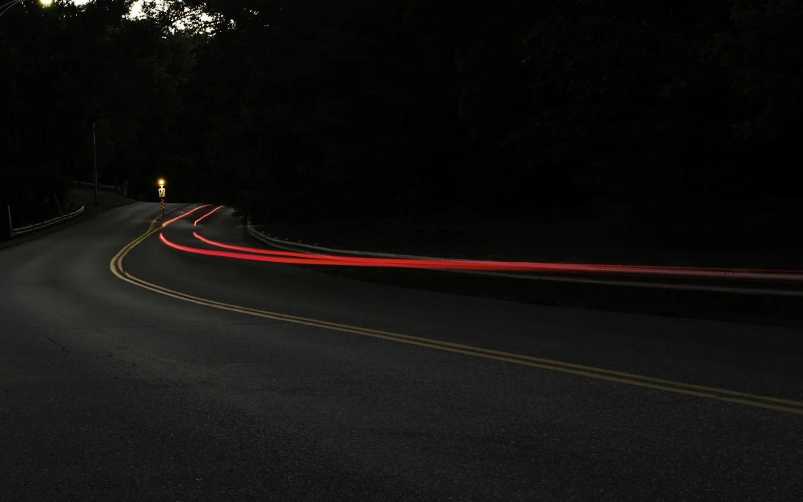 A road curving through trees at night, with red tail-light trails stretching around the bend.